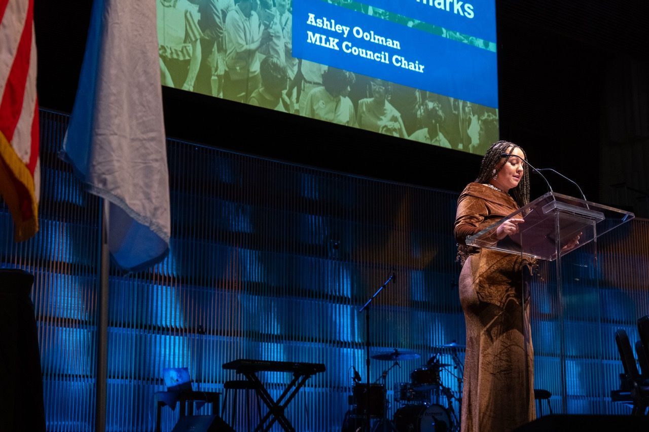 A woman with braided hair stands at a clear podium speaking on stage with a blue-lit background. Two flags are partially visible on the left. A projection screen above displays the text "Ashley Oolman MLK Council Chair." Musical instruments, including a keyboard and drums, are visible in the background.